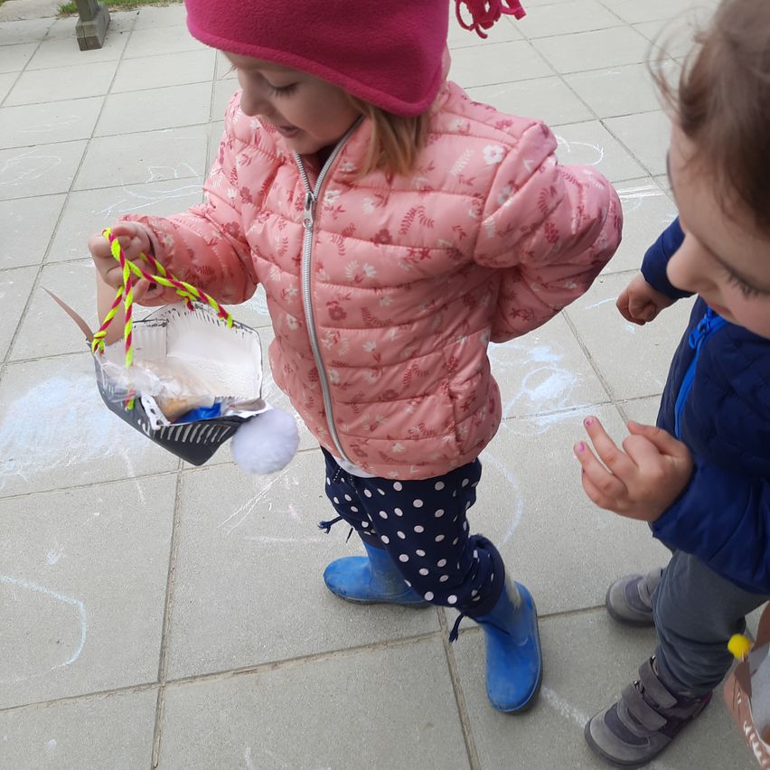Ein Mädchen in einer rosa Jacke und Mütze hält einen Papierkorb mit einem Pompon, während ein junges Kind zusieht.
