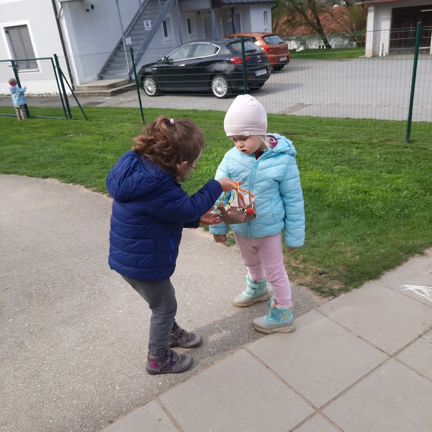 Zwei Mädchen stehen auf einem Bürgersteig, eines hält einen kleinen Korb. Dahinter befindet sich ein Rasenbereich mit einem Zaun. Vor einem Gebäude mit einer Treppe sind Autos geparkt.