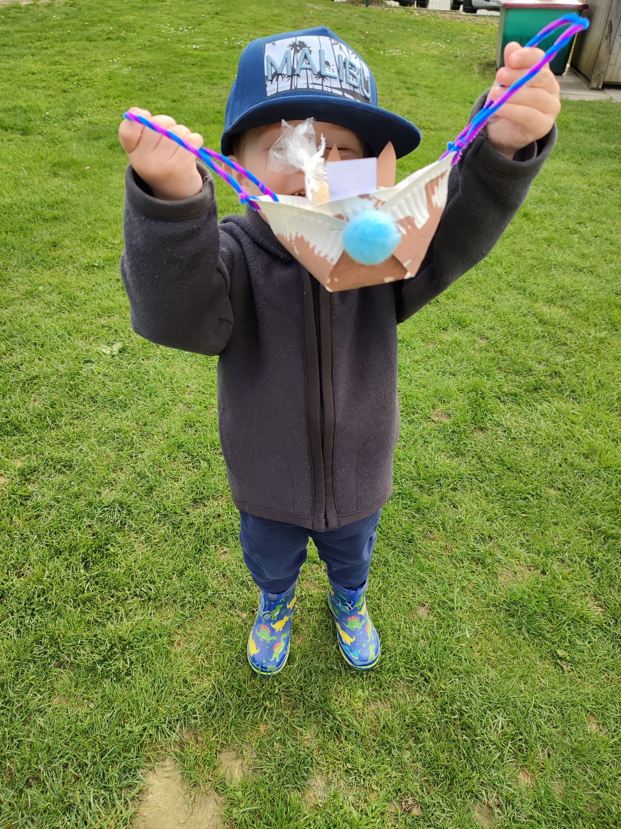 Ein kleiner Junge in einem schwarzen Jacken und einer blauen Mütze hält eine selbstgemachte Maske aus Papier und Schnur. Die Maske hat einen bunten Pompon und ist über seinem Gesicht positioniert.