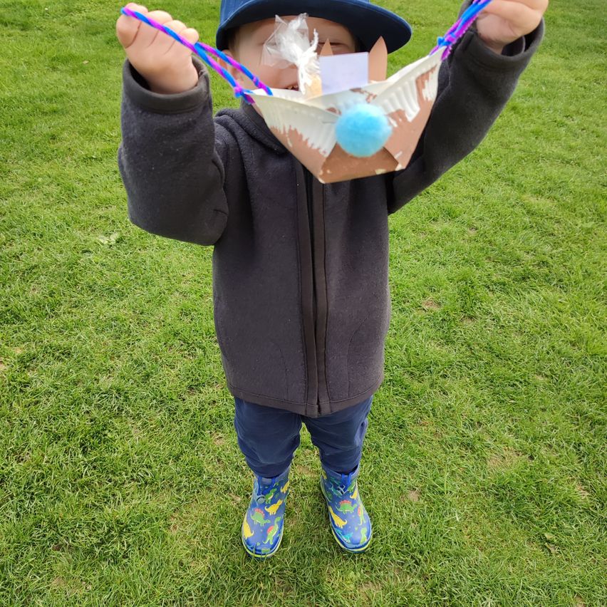 Ein kleiner Junge in einem schwarzen Jacken und einer blauen Mütze hält eine selbstgemachte Maske aus Papier und Schnur. Die Maske hat einen bunten Pompon und ist über seinem Gesicht positioniert.