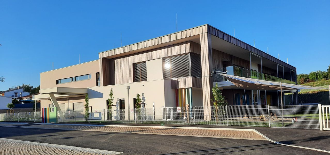 Ein modernes Gebäude mit Holzfassade und Glasfenstern steht an einem sonnigen Tag. Das Gebäude verfügt über eine bunt gestreifte Wand und einen Balkon. Ein Zaun umgibt den vorderen Bereich.