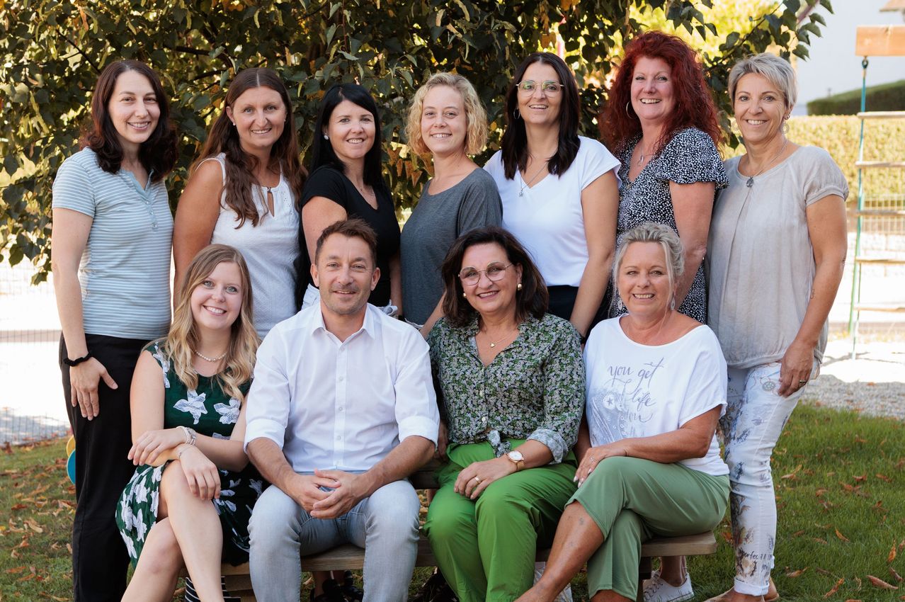 Eine Gruppe von Frauen und zwei Männern lächelt und posiert für ein Foto im Garten. Die Frauen tragen Freizeitkleidung. Der Mann in der Mitte trägt ein weißes Hemd.