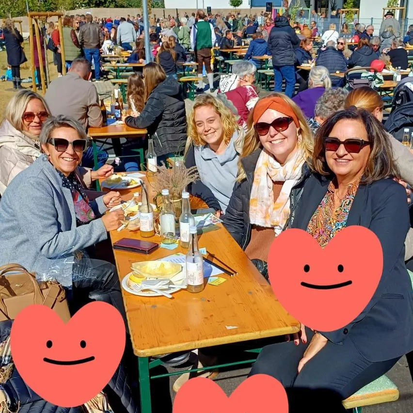 Eine Gruppe von Frauen sitzt an einem Tisch in einem Restaurant im Freien. Sie tragen Sonnenbrillen und Schals. Auf dem Tisch stehen Teller mit Essen und Flaschen. Im Hintergrund sitzen viele Menschen an anderen Tischen.