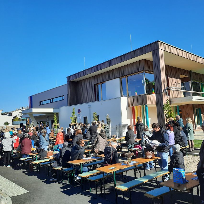Eine Menschenmenge versammelt sich vor einem modernen Gebäude mit Glasfenstern und sitzt an Tischen unter einem klaren blauen Himmel.