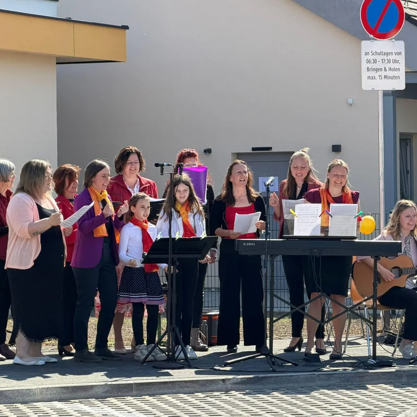 Eine Gruppe von Erwachsenen und Kindern singt auf der Straße vor einem Gebäude. Eine Frau spielt Gitarre, während andere Papiere halten.