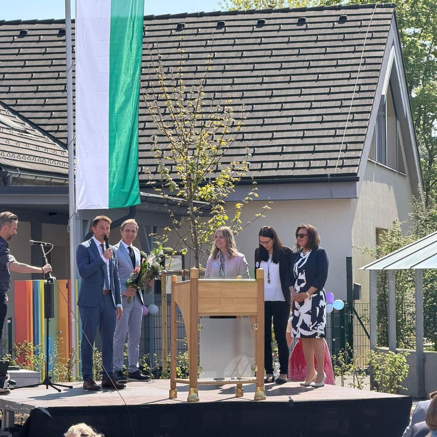 Mehrere Personen stehen auf einer Bühne vor einem Haus. Ein Mann hält ein Mikrofon und einen Blumenstrauß. Ein Baum steht auf der Bühne. Eine Fahne weht hinter ihnen.