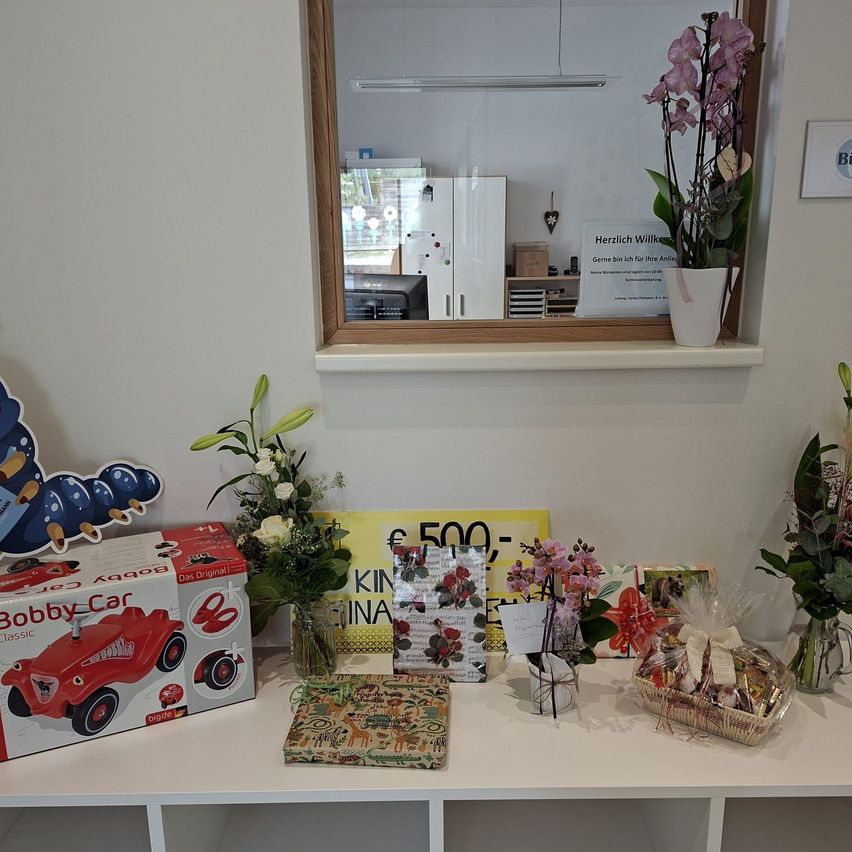 Ein Regal mit Geschenken und Blumen. Ein Bobby Car Spielzeug, ein Buch, ein Blumenstrauß, ein Geschenkkorb und ein Schild mit Preisschild. Ein Spiegel reflektiert einen Kühlschrank und ein Schild.
