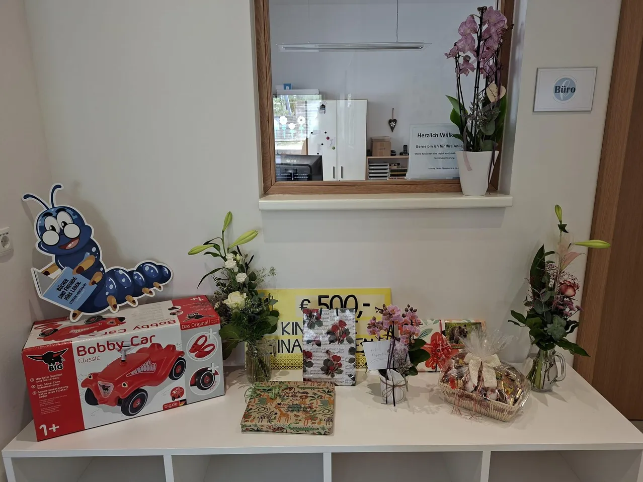Ein Regal mit Geschenken und Blumen. Ein Bobby Car Spielzeug, ein Buch, ein Blumenstrauß, ein Geschenkkorb und ein Schild mit Preisschild. Ein Spiegel reflektiert einen Kühlschrank und ein Schild.