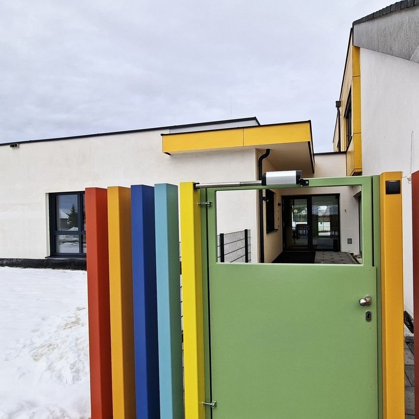 Ein farbenfrohes Haus mit einem Regenbogen-farbigen Zaun und einer grünen Tür mit Schloss und Griff, in einem verschneiten Gebiet gelegen.