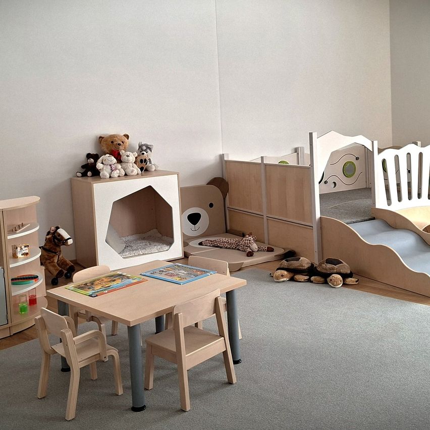 Ein gemütliches Kinderzimmer mit hellen Holzmöbeln, einem Tisch mit Büchern und einem Bett mit Hundehütten-Design. Gefüllte Tiere stehen auf Regalen und im ganzen Raum.