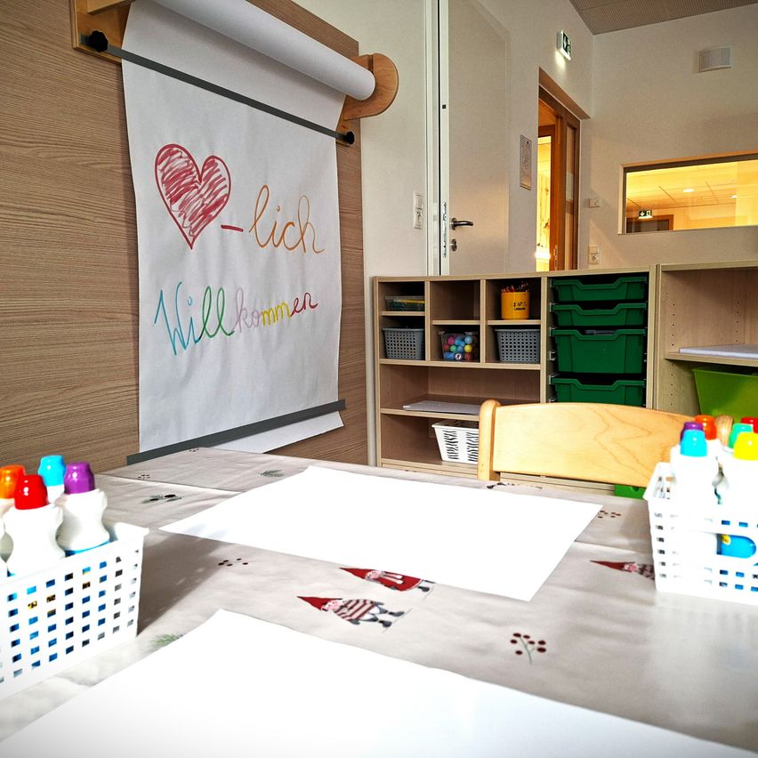 Ein Raum mit einem Tisch, der mit weißem Tuch und Papier bedeckt ist, Aufbewahrungsboxen, einem Stuhl und Regalen. Ein Poster mit einem Herz und Text ist an der Wand montiert.