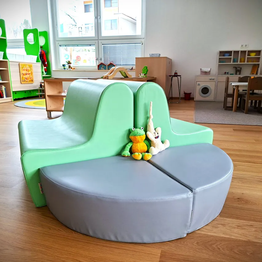 Ein Raum mit Holzboden, einem grünen gebogenen Sofa und Kuscheltieren darauf. Dahinter befinden sich Regale, ein Tisch und Stühle. Rechts gibt es eine Waschmaschine und einen Schrank.
