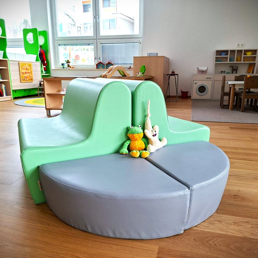 Ein Raum mit Holzboden, einem grünen gebogenen Sofa und Kuscheltieren darauf. Dahinter befinden sich Regale, ein Tisch und Stühle. Rechts gibt es eine Waschmaschine und einen Schrank.