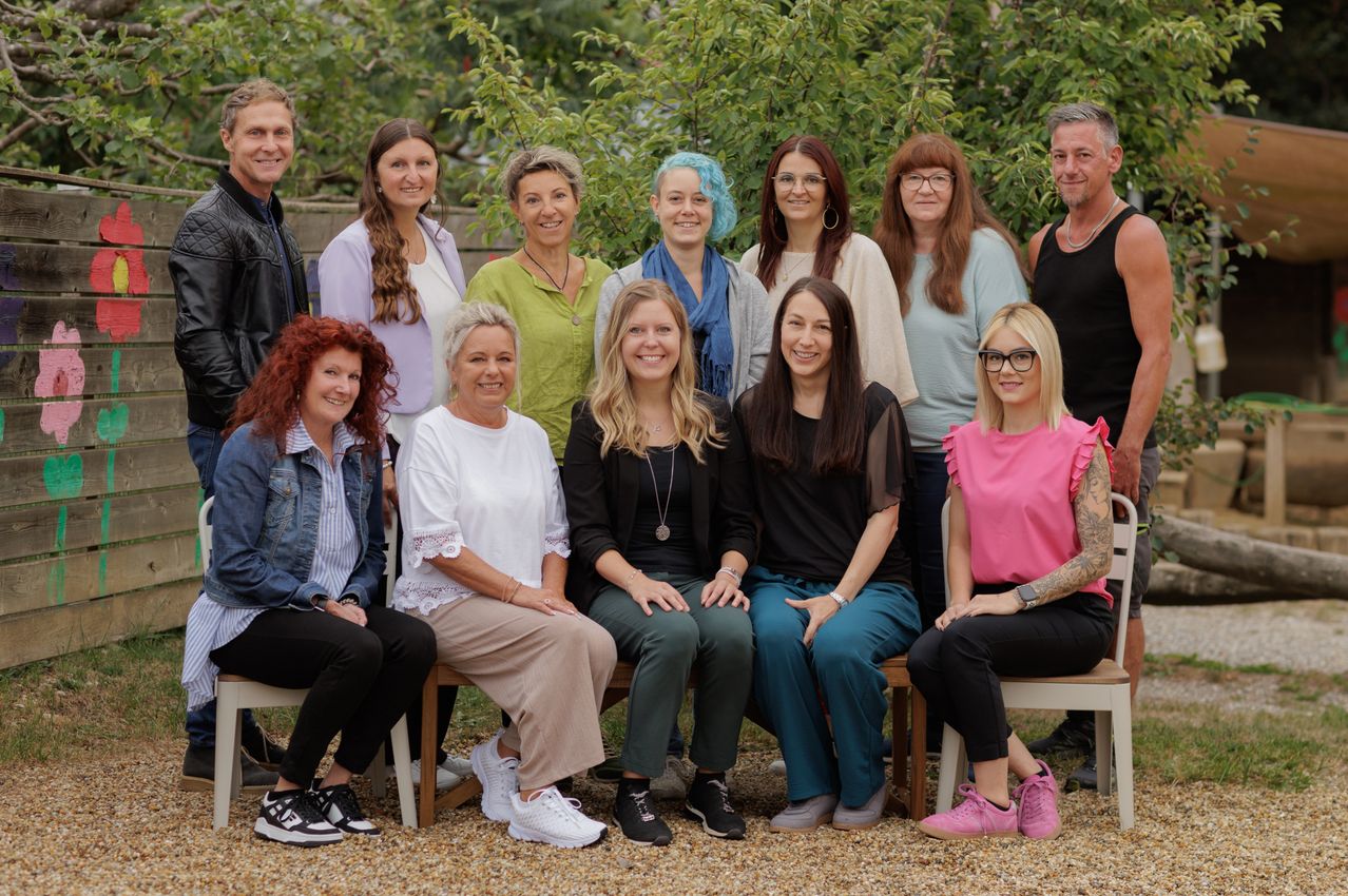 Eine Gruppe von Menschen, sowohl Männer als auch Frauen, sitzen auf Stühlen in einer Außenumgebung. Sie lächeln und scheinen für ein Foto zu posieren. Der Hintergrund zeigt viel Grün.