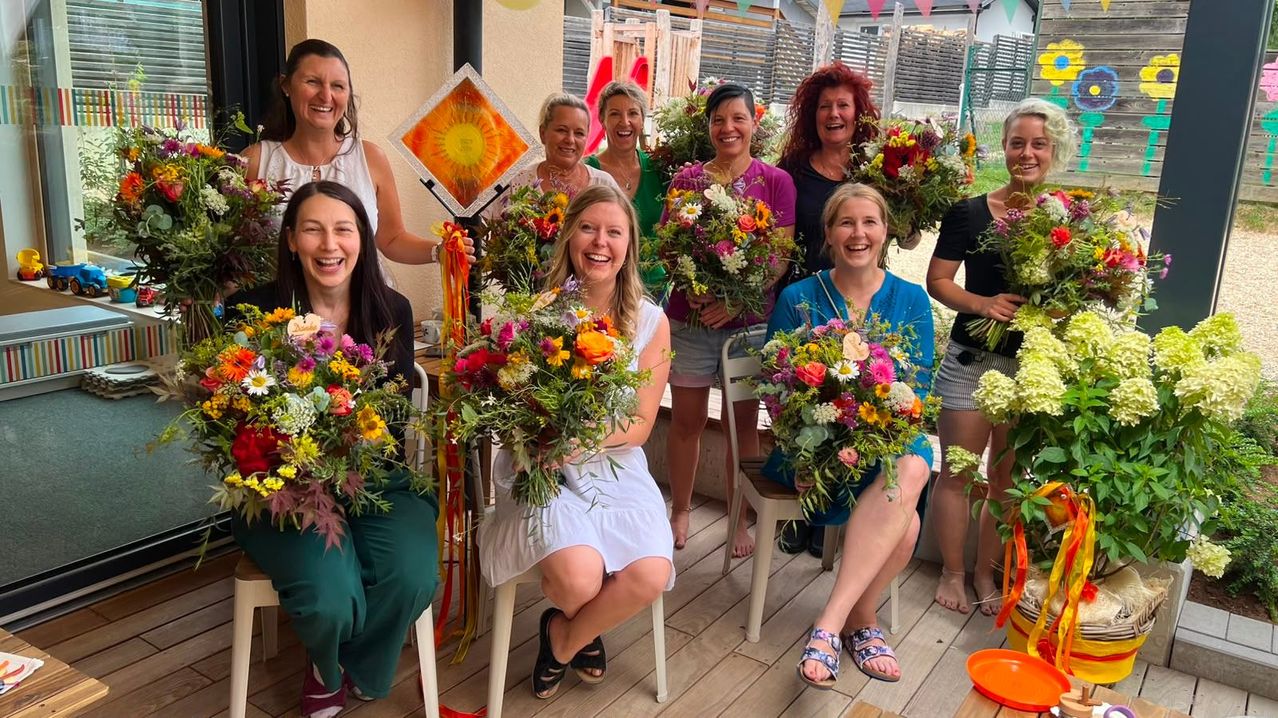 Eine Gruppe von Frauen lächelt und posiert für ein Foto, während sie Blumensträuße halten. Sie sitzen auf Stühlen auf einer Holzterrasse.