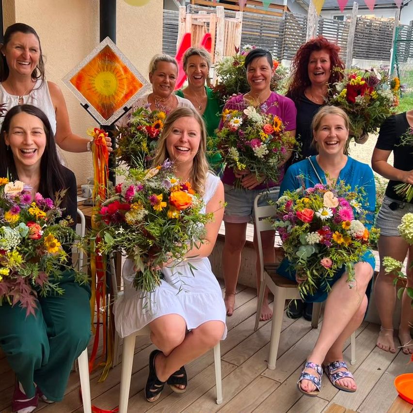 Eine Gruppe von Frauen lächelt und posiert für ein Foto, während sie Blumensträuße halten. Sie sitzen auf Stühlen auf einer Holzterrasse.