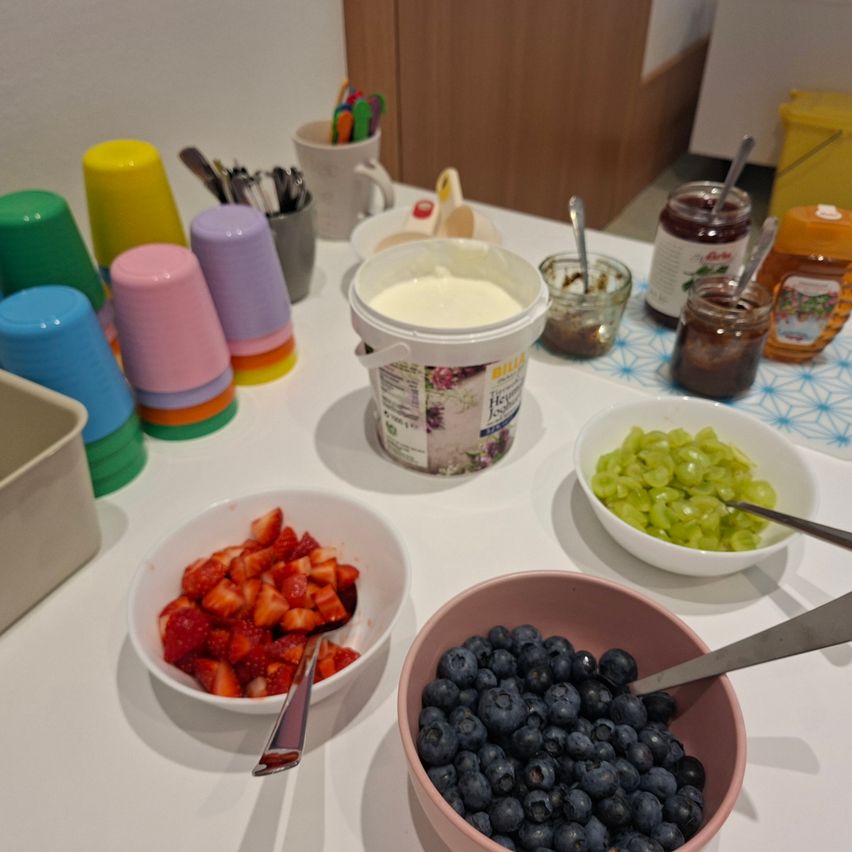 Ein Tisch mit Joghurt, Obst und Beilagen. Schüsseln mit Erdbeeren und Blaubeeren, ein Behälter mit Joghurt und Gläser mit Marmelade und Honig sind auf einer weißen Tischdecke angeordnet. Bunte Tassen und Besteck sind ebenfalls vorhanden.
