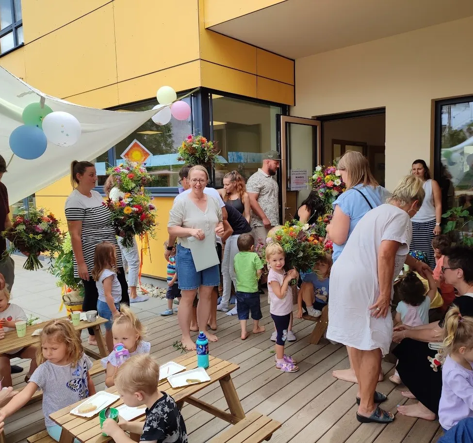Eine Gruppe von Erwachsenen und Kindern versammelt sich auf einer Holzterrasse, einige halten Blumen, mit Luftballons und einem Gebäude im Hintergrund.