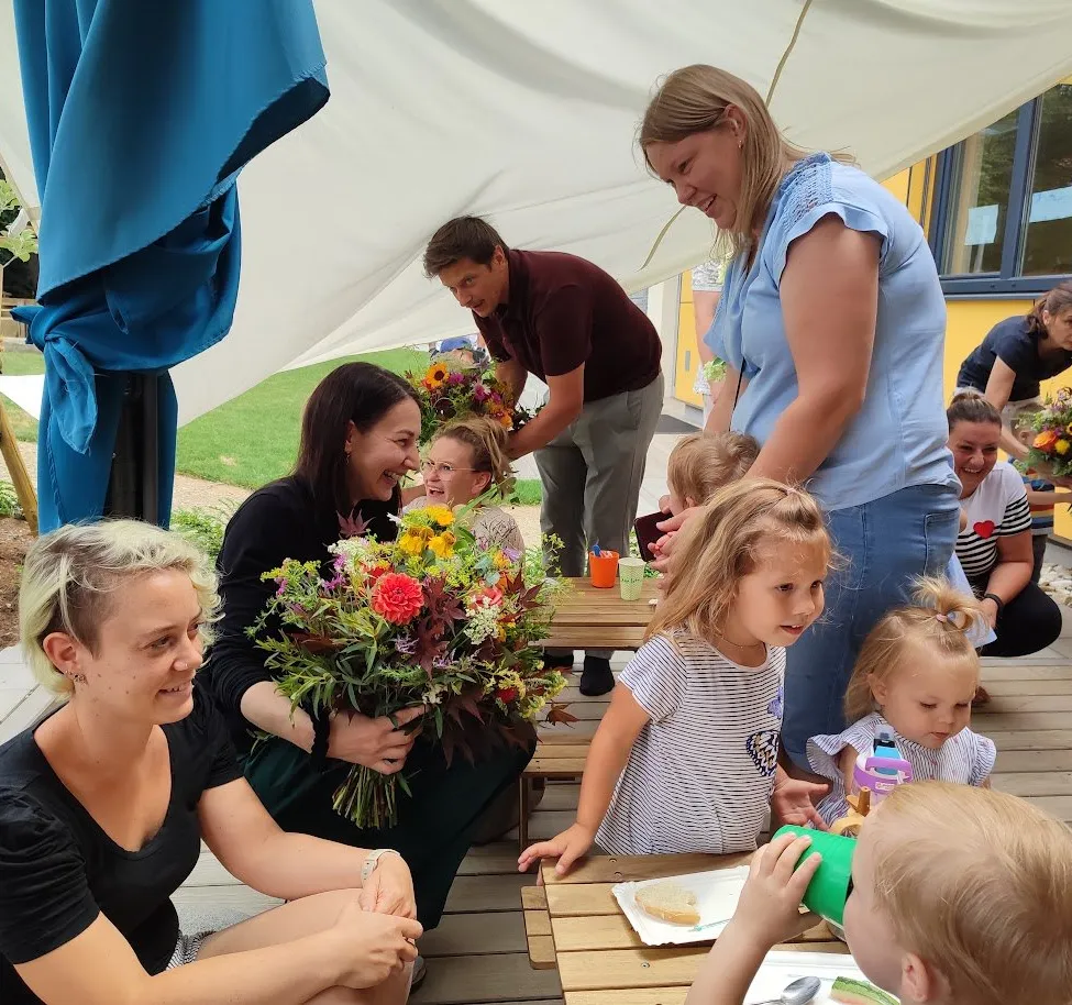 Eine Gruppe von Erwachsenen und Kindern versammelt sich unter einem Zelt, wobei eine Frau einen großen Blumenstrauß hält. Die Kinder sitzen um einen Tisch herum, und ein Kind isst.