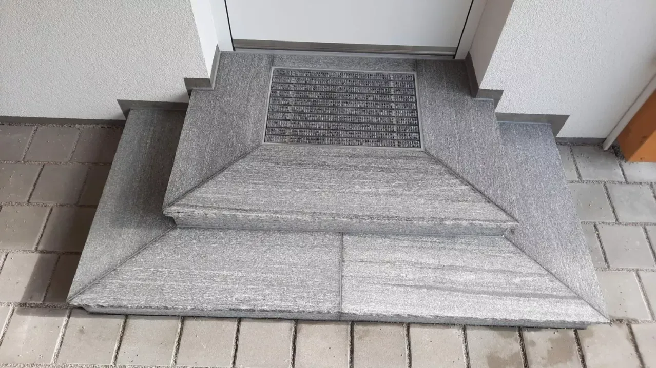 Eine Nahaufnahme einer Betonstufe mit einem silbernen Gitter darüber, neben einer weißen Wand.