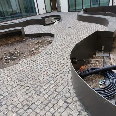 Eine Baustelle mit einer gebogenen schwarzen Struktur und Kopfsteinpflaster. Schutt und ein Schlauch sind vorhanden. Glasfassadengebäude im Hintergrund.