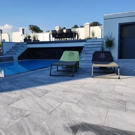 An outdoor pool area with two lounge chairs by the pool, surrounded by stone tiles and steps leading to a deck with a table and chairs.