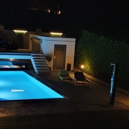 A swimming pool at night, illuminated with blue lights. Two lounge chairs are placed on the right side, and a small plant is in front of the door.