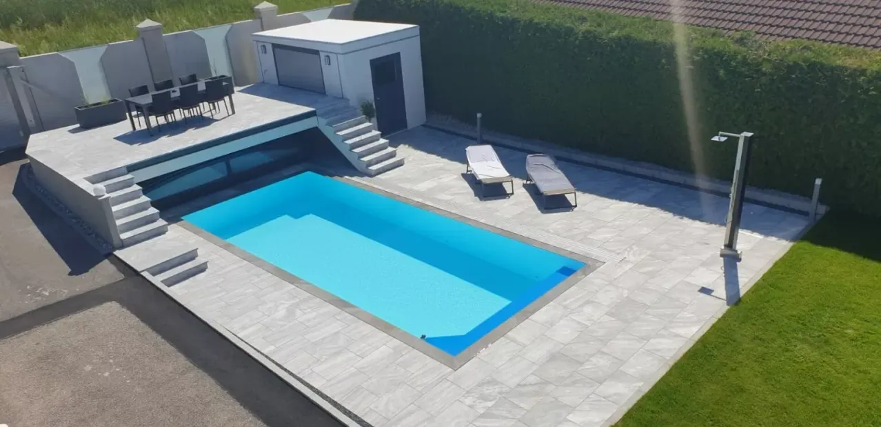 An aerial view of a swimming pool surrounded by stone tiles, with two lounge chairs nearby, a white shed, and a wooden deck.
