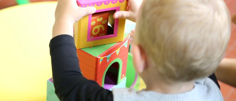 Ein junger Junge spielt mit einem farbenfrohen Blockturm und versucht, eine rosafarbene Box oben drauf zu balancieren. Er steht auf einer gelben Oberfläche.