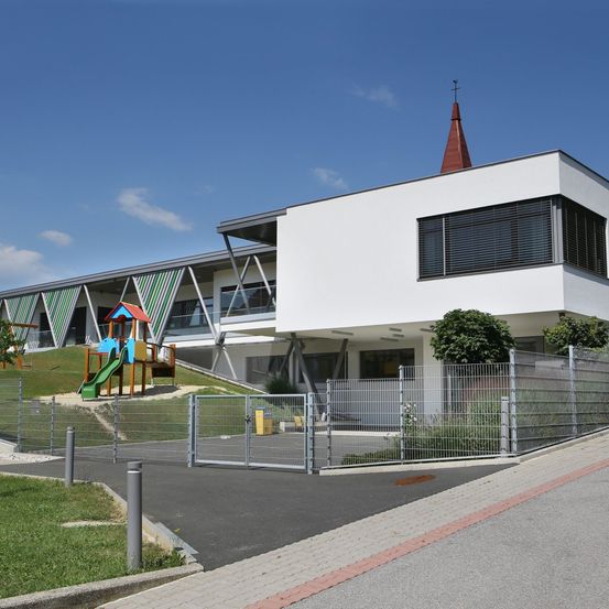 Ein modernes weißes Gebäude mit einem roten Turm steht in der Nähe eines Spielplatzes und eines eingezäunten Bereichs. Das Gebäude hat große Fenster und ein geneigtes Dach.