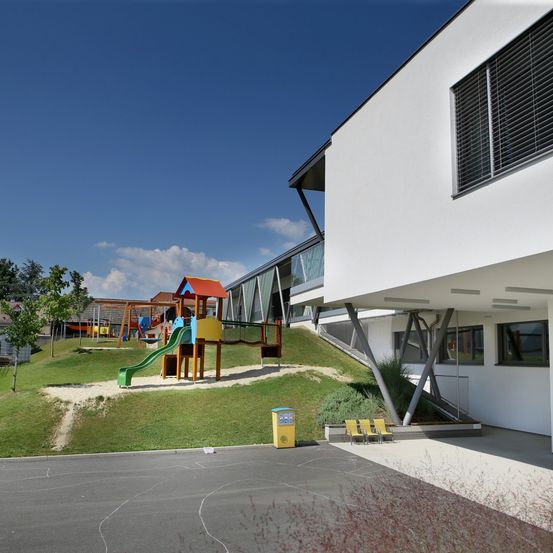 Ein modernes Gebäude mit einem Spielplatz auf einem Rasenhang. Der Spielplatz hat eine Rutsche und ein Klettergerüst. Das Gebäude hat Glasfenster und eine Metallkonstruktion.
