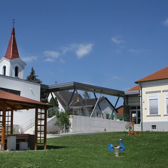 Eine Kirche mit einem Turm steht neben einem modernen Gebäude. Die Kirche hat ein rotes Dach und eine weiße Fassade. In der Nähe befindet sich ein Pavillon und ein Spielplatz. Das Gebiet ist von Gras und einer Betonmauer umgeben.