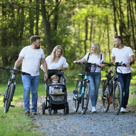 Eine Familie mit fünf Personen geht durch einen bewaldeten Bereich auf einem Schotterweg, wobei zwei Erwachsene Fahrräder fahren und ein Baby in einem Kinderwagen ist.