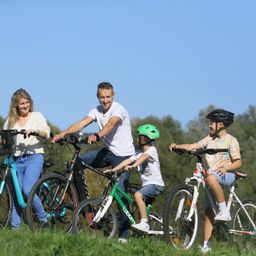 Eine vierköpfige Familie, zwei Erwachsene und zwei Kinder, posiert für ein Foto auf einem Grasfeld mit ihren Fahrrädern. Die Kinder tragen Helme. Der Himmel ist klar und blau.