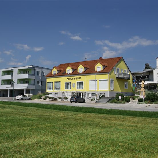 Ein gelbes Gebäude mit rotem Dach und weißen Fenstern, beschriftet mit Gemeindeamt, steht vor einem grasigen Feld. Ein Auto ist davor geparkt.