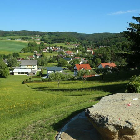 Ein Dorf umgeben von grünen Hügeln, mit Häusern in verschiedenen Farben, einer Kirche und einem großen Felsen im Vordergrund.