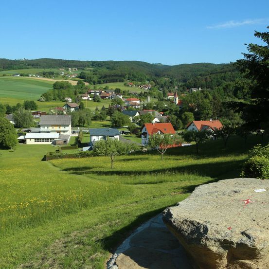 Ein Dorf umgeben von grünen Hügeln, mit Häusern in verschiedenen Farben, einer Kirche und einem großen Felsen im Vordergrund.