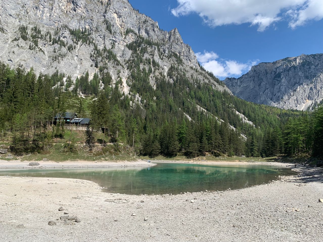 Ein Bergsee mit grünem Wasser und Felsen am Ufer. Ein Haus steht am Berghang.