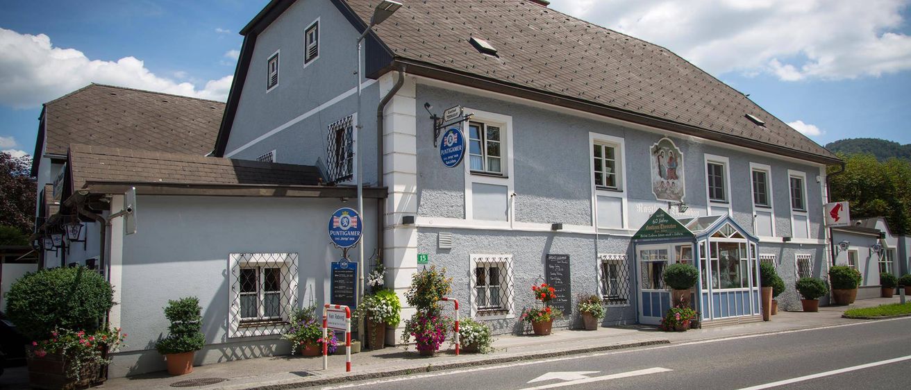 Ein traditionelles Haus mit einem braunen Dach und mehreren Fenstern. Ein blaues Schild mit Text ist an dem Pfosten auf der linken Seite befestigt. Das Haus ist von Topfpflanzen und Blumen umgeben.