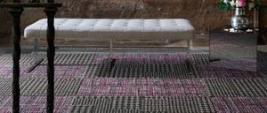 Eine Bank steht auf einem karierten Teppich vor einer strukturierten Steinwand mit einer Blumenvase auf einem Tisch.