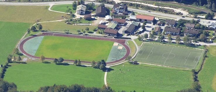 Ein Luftbild eines Sportkomplexes mit einem Fußballfeld, einer Laufbahn und mehreren Häusern im Hintergrund.