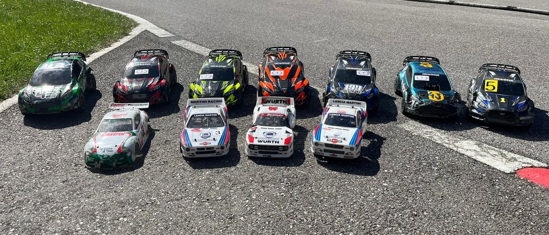 Sieben Spielzeugautos sind auf einer Rennstrecke aufgereiht. Drei sind weiß, zwei sind schwarz, eines ist blau und eines ist orange. Alle sind Rennmodelle mit verschiedenen Sponsoren und Nummern.