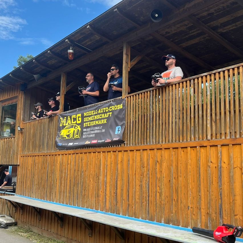 Mehrere Männer stehen auf einem Balkon und blicken auf eine Rennstrecke, halten Fernbedienungen in der Hand. Sie scheinen an einem Modellauto-Cross-Event teilzunehmen.