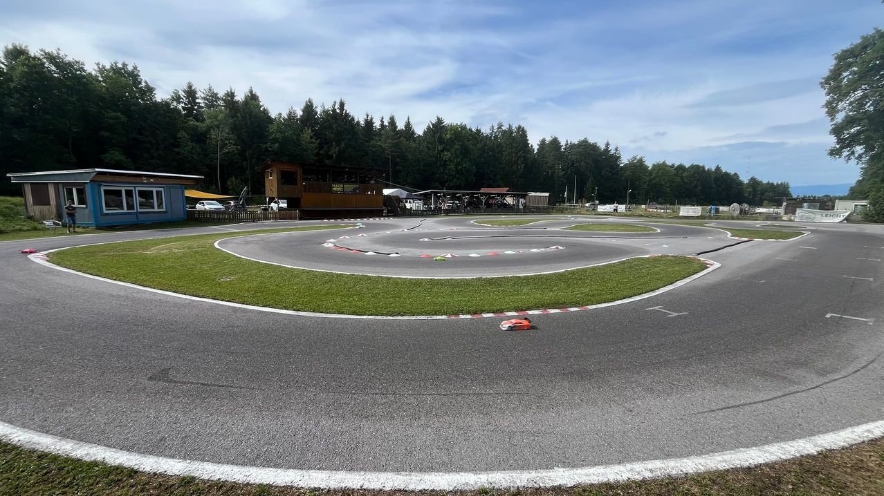 Eine Rennstrecke mit einem roten Auto auf der Strecke. Grüner Rasen umgibt die Strecke, und ein Gebäude ist in der Ferne.