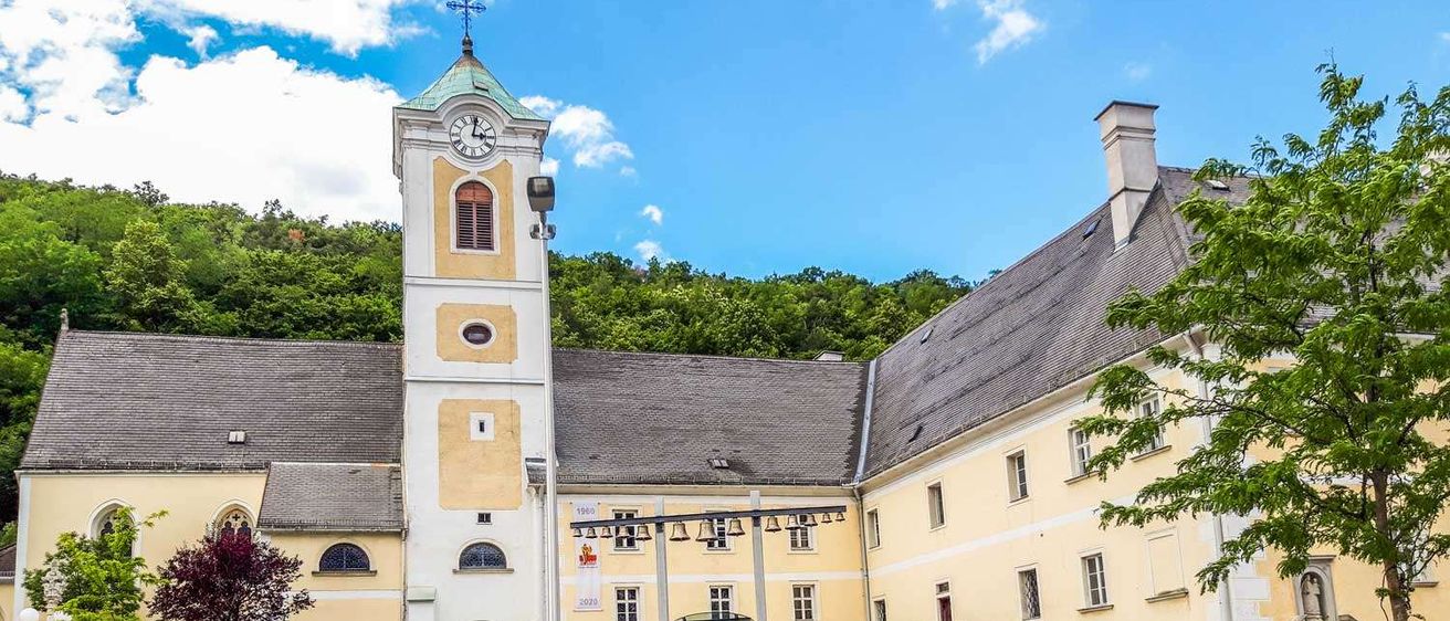 Die Vorderseite eines großen Gebäudes mit einem Glockenturm und einem Weg, der zum Eingang führt. Davor stehen Topfpflanzen und Bäume.