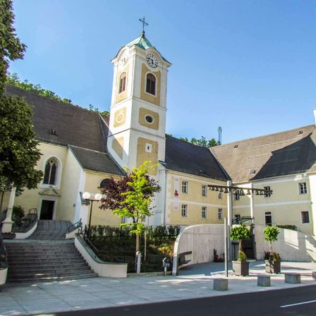 Die Vorderseite einer Kirche mit einem Glockenturm und Stufen, die zum Eingang führen. Das Gebäude ist weiß und hat ein graues Dach.