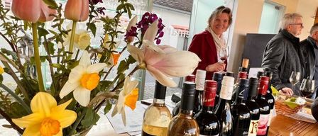 Eine Frau lächelt und hält ein Weinglas in einem Raum mit Glasfenster. Auf dem Tisch steht eine Vase mit Blumen und mehrere Flaschen Wein. Dahinter ist eine verschwommene Ansicht eines Gebäudes.