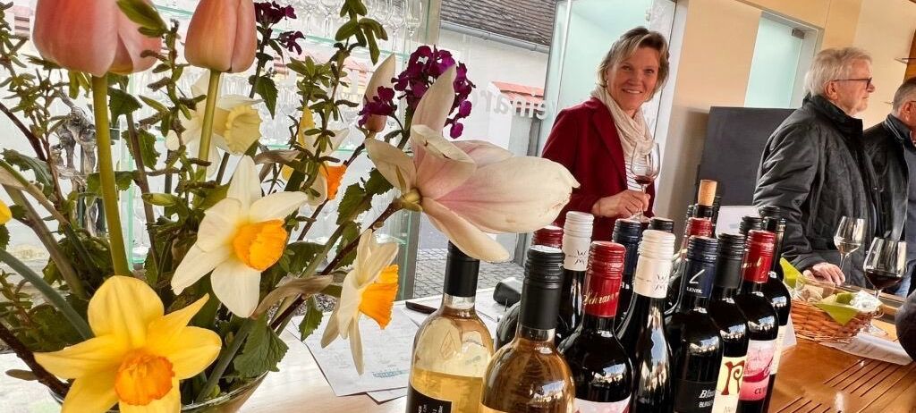 Eine Frau lächelt und hält ein Weinglas in einem Raum mit Glasfenster. Auf dem Tisch steht eine Vase mit Blumen und mehrere Flaschen Wein. Dahinter ist eine verschwommene Ansicht eines Gebäudes.