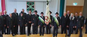 Eine Gruppe von Männern in Uniformen steht mit einer Flagge zusammen und lächelt für ein Foto. Sie tragen Hüte, Schärpen und Krawatten. Hinter ihnen ist eine Wand mit einem Schild.