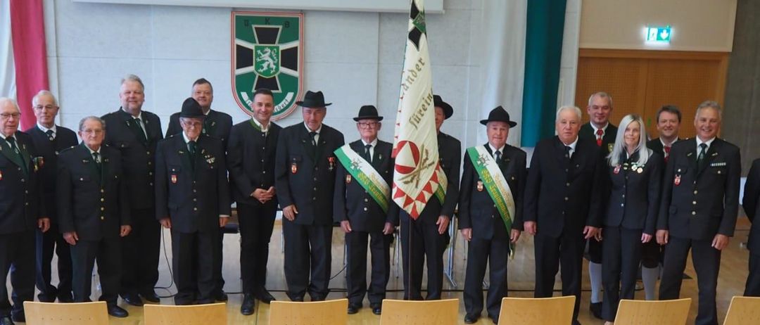 Eine Gruppe von Männern in Uniformen steht mit einer Flagge zusammen und lächelt für ein Foto. Sie tragen Hüte, Schärpen und Krawatten. Hinter ihnen ist eine Wand mit einem Schild.